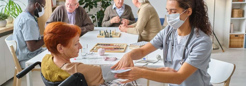 Nurse measuring the pressure to senior woman who using wheelchair in the nursing home with other senior people playing chess in the background