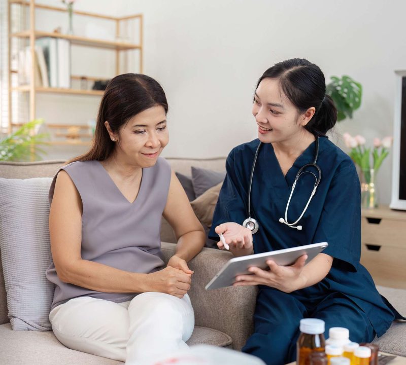 A healthcare professional in blue scrubs consults with a patient, using a tablet to discuss medical information in a cozy home environment.