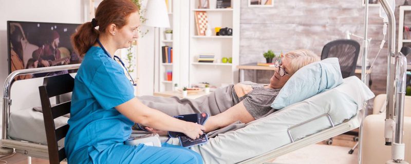 Female doctor checking senior woman blood pressure lying in bed. Nursing home.